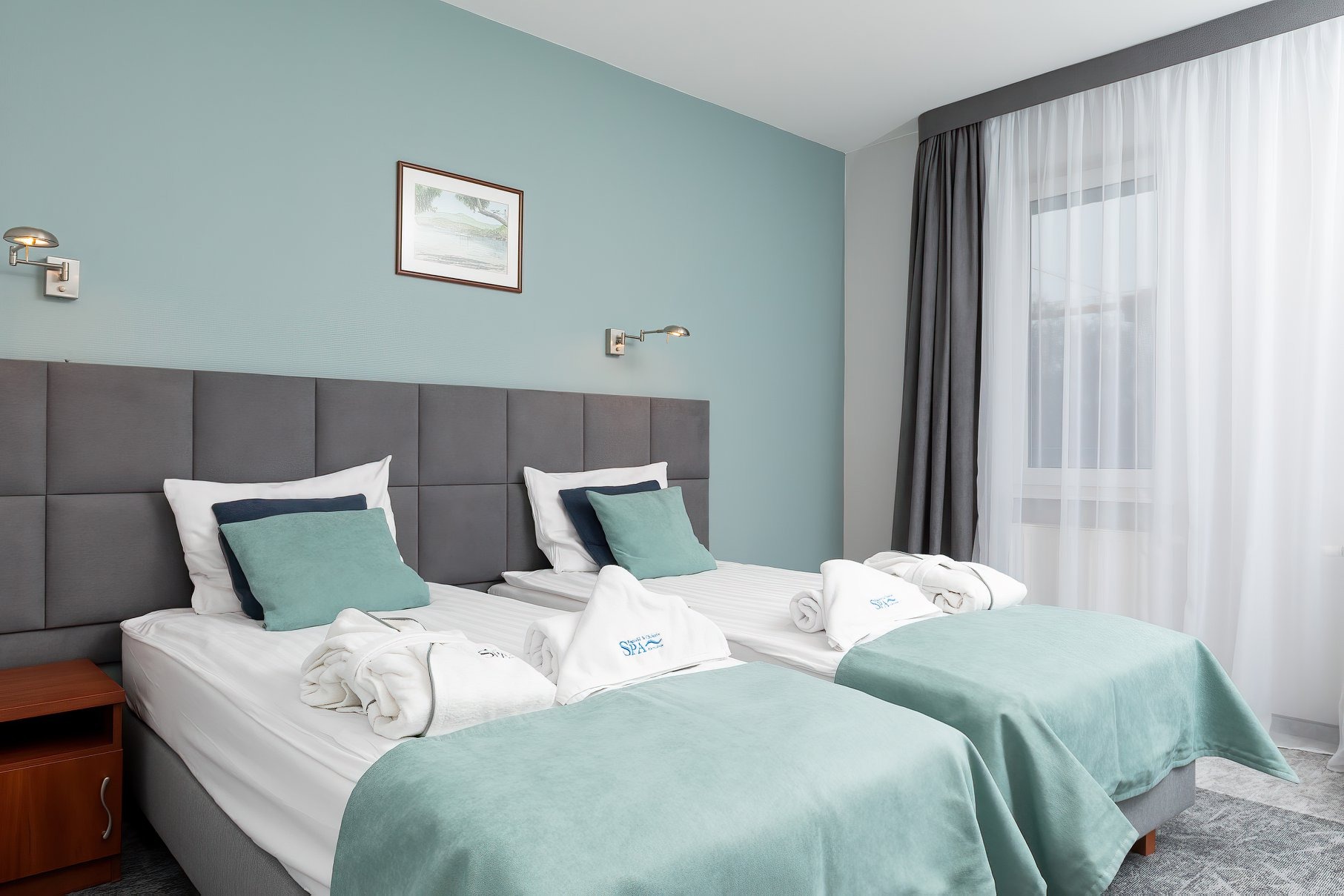 Pokój hotelowy z dwoma łóżkami, szarą tapicerowaną ścianą, turkusowymi akcentami i ręcznikami z logo SPA.