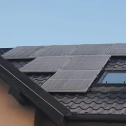 Panele słoneczne na ciemnym dachu z dachówką ceramiczną, widoczne okno dachowe, lekko zachmurzone niebo.