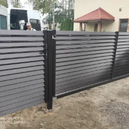 Nowoczesna, przesuwna brama wjazdowa z poziomych profili w kolorze ciemnego brązu, widoczna na tle jasnego domu i białego samochodu dostawczego z ekipą montażową.