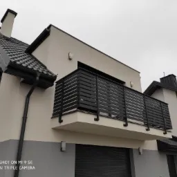 Nowoczesny balkon z czarną, poziomą balustradą na tle dwukolorowej elewacji domu z szarą bramą garażową.