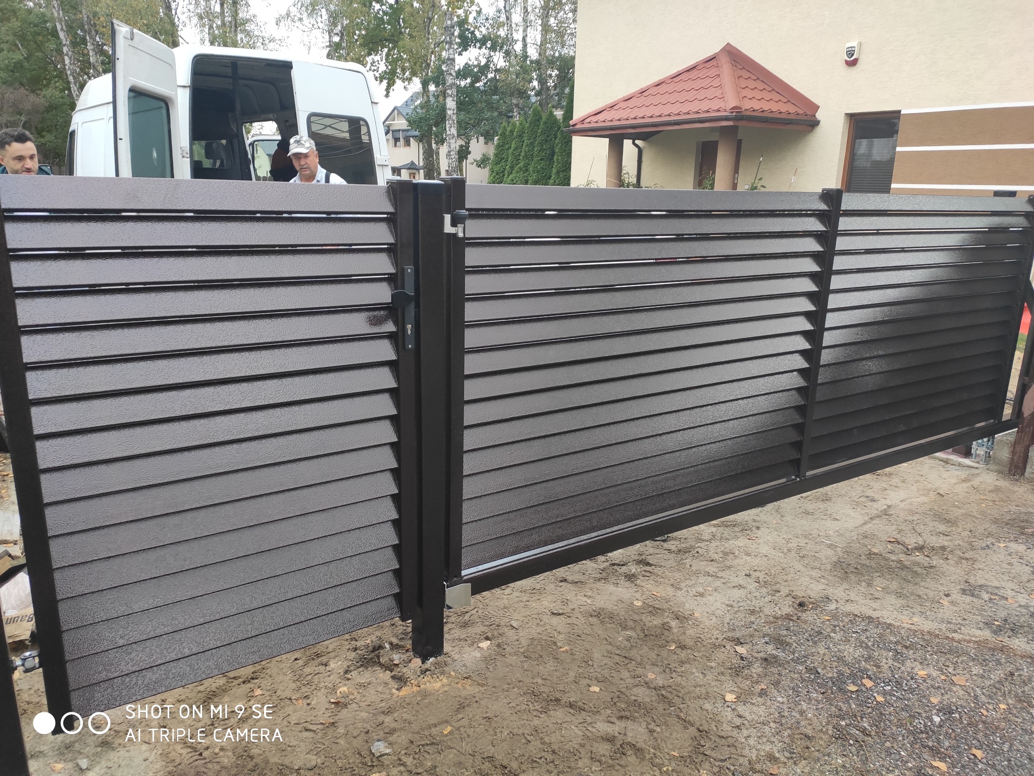 Nowoczesna, przesuwna brama wjazdowa z poziomych profili w kolorze ciemnego brązu, widoczna na tle jasnego domu i białego samochodu dostawczego z ekipą montażową.