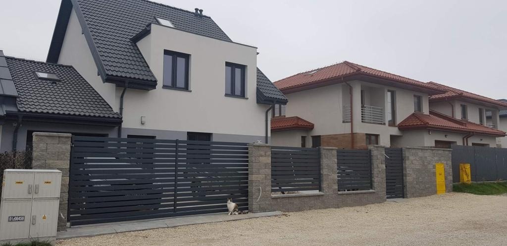 Nowoczesne ogrodzenie poziome z szarych paneli na podmurówce z szarej kostki, widok na dwa domy jednorodzinne w tle, kot siedzi przed ogrodzeniem.