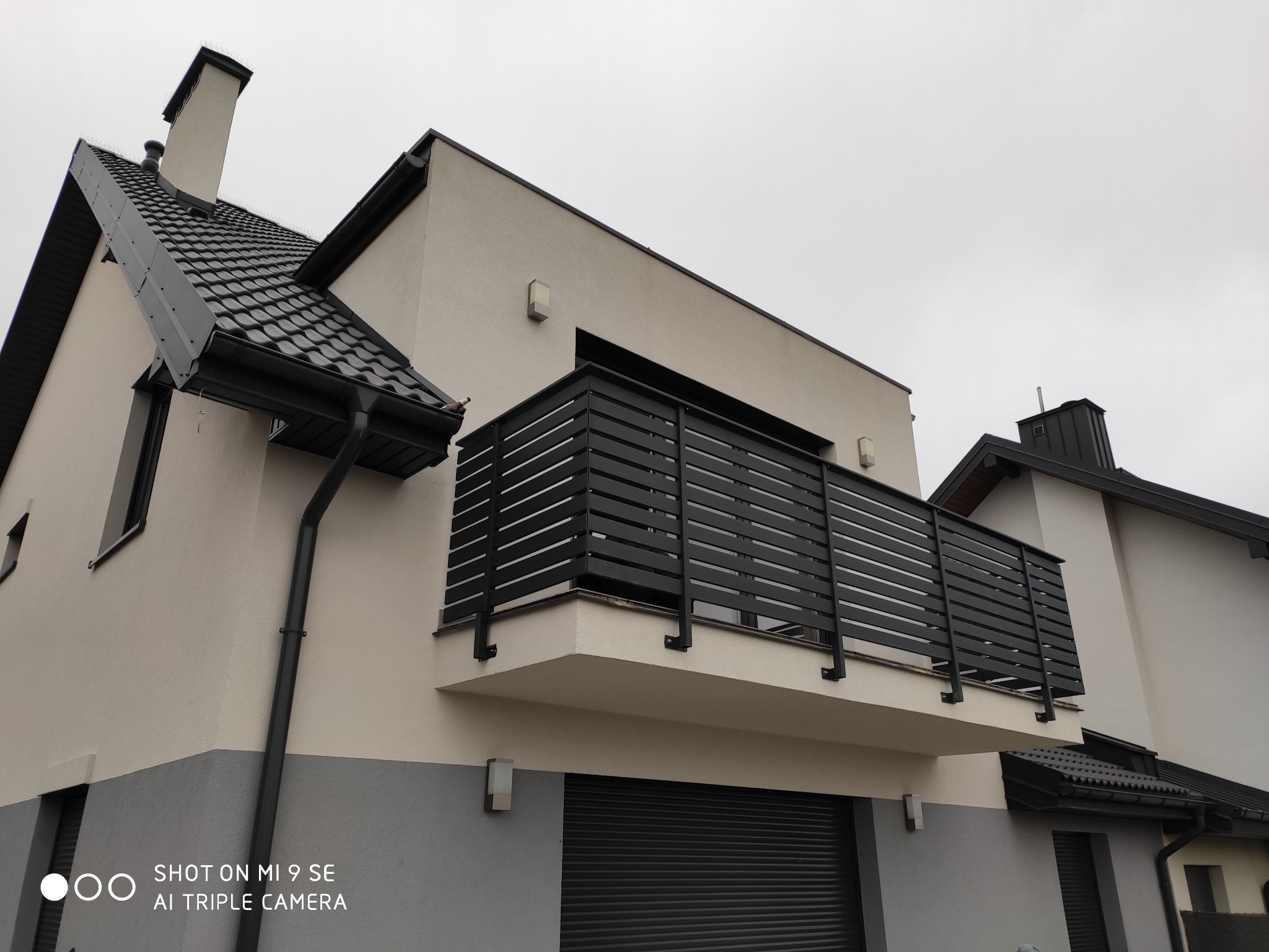 Nowoczesny balkon z czarną, poziomą balustradą na tle dwukolorowej elewacji domu z szarą bramą garażową.