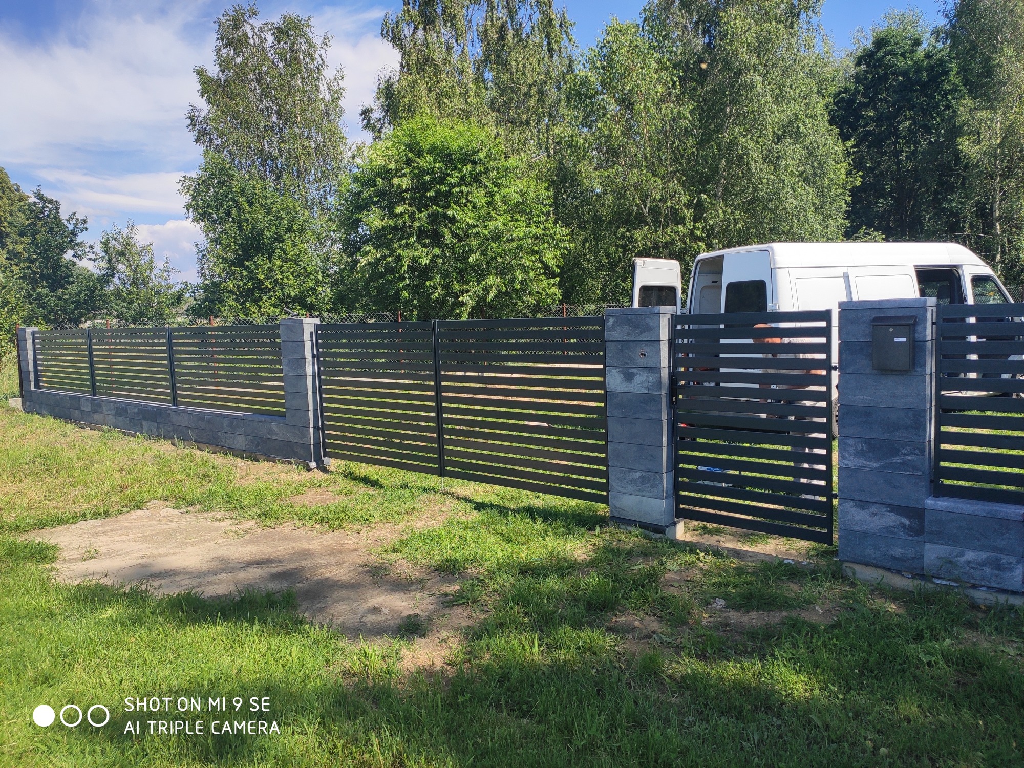 Nowoczesne ogrodzenie panelowe z poziomymi przęsłami i słupkami z szarego kamienia łupanego, brama wjazdowa z czarną skrzynką na listy, biały samochód dostawczy w tle, zielony trawnik.