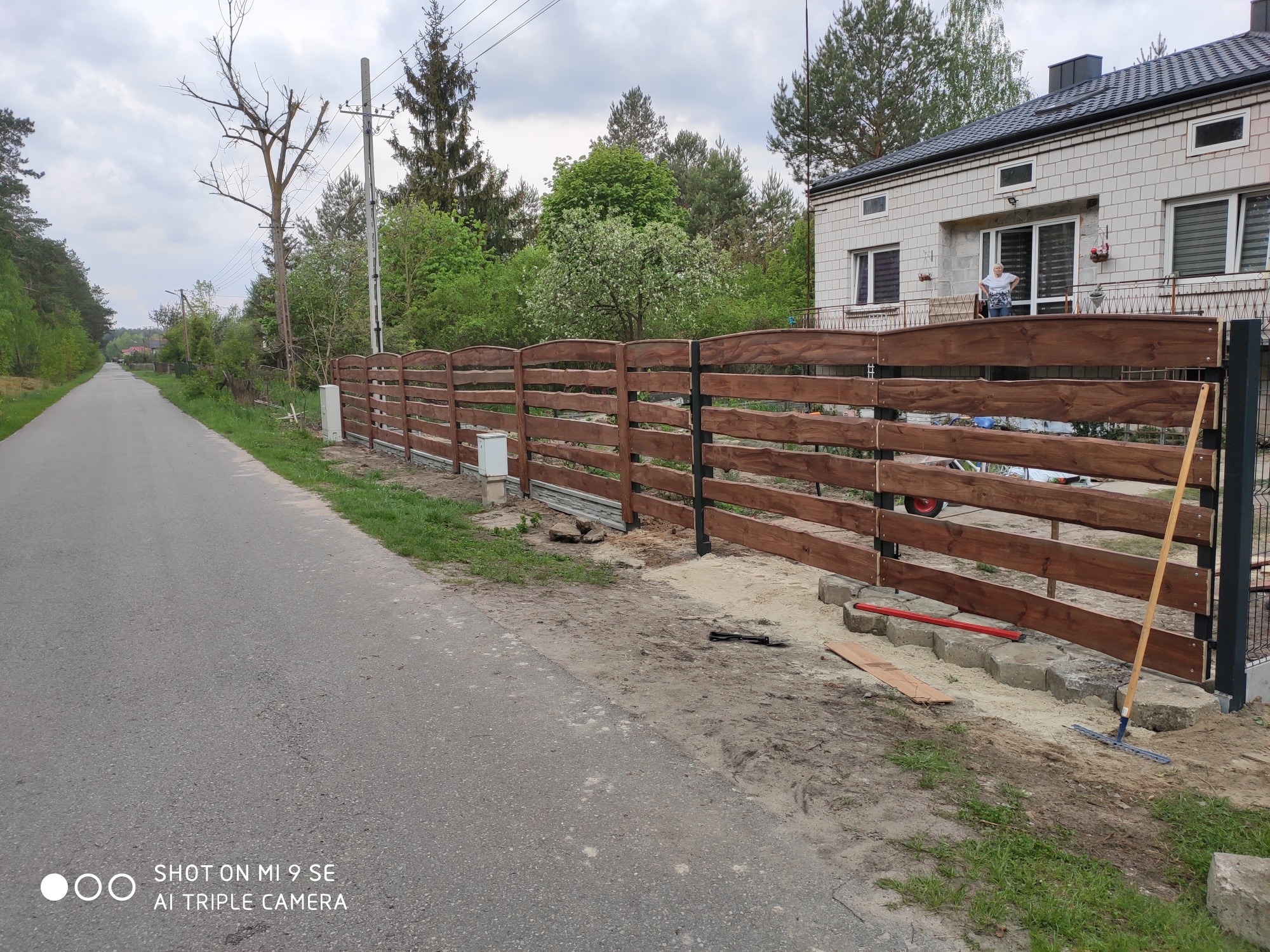 Nowoczesne ogrodzenie z poziomych desek w kolorze brązu, zamontowane na czarnych metalowych słupkach, widok z ulicy na tle domu i zieleni, na ziemi widoczne narzędzia i materiały budowlane.