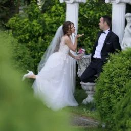 Panna młoda w koronkowej sukni ślubnej z welonem, posyła całusa w stronę pana młodego w smokingu, który siedzi na kamiennej balustradzie w ogrodzie z kolumnami i rzeźbą.