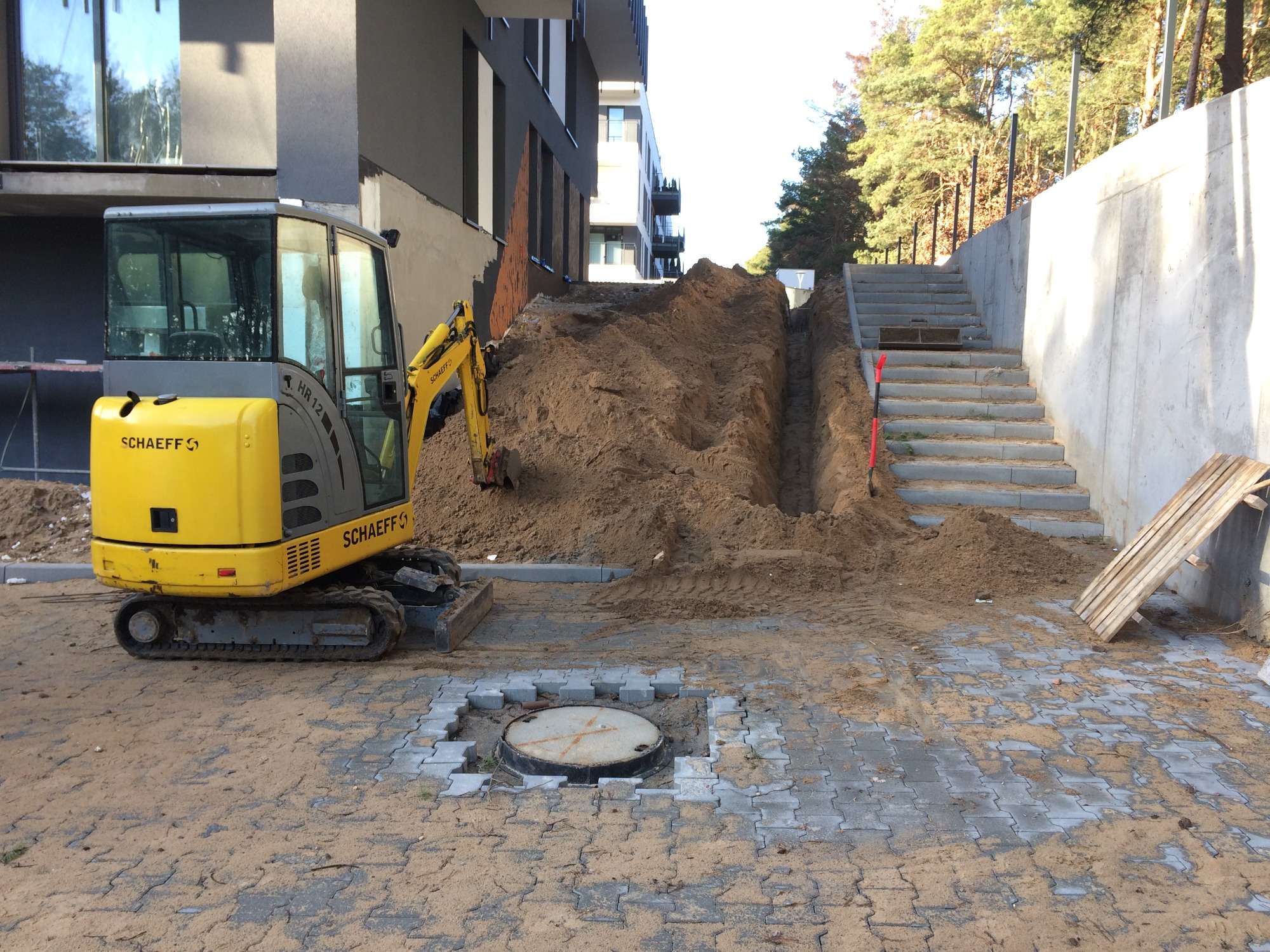 Żółta mini koparka Schaeff przy wykopie wąskiego rowu w piaszczystej ziemi obok nowo budowanego budynku apartamentowego i betonowych schodów.