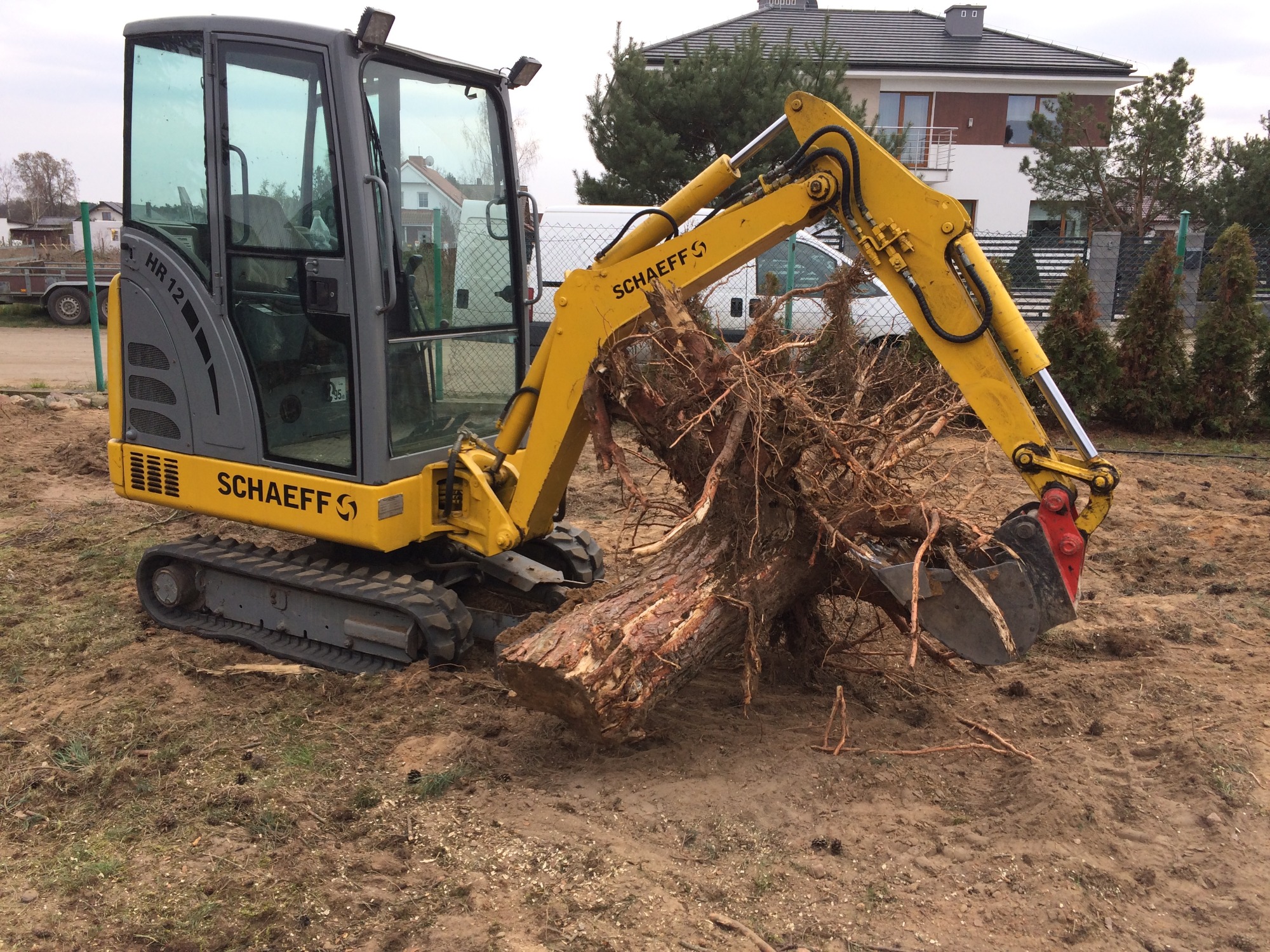 Żółto-szara minikoparka marki Schaeff chwyta pniem z korzeniami na placu budowy, w tle dom jednorodzinny i biały bus.
