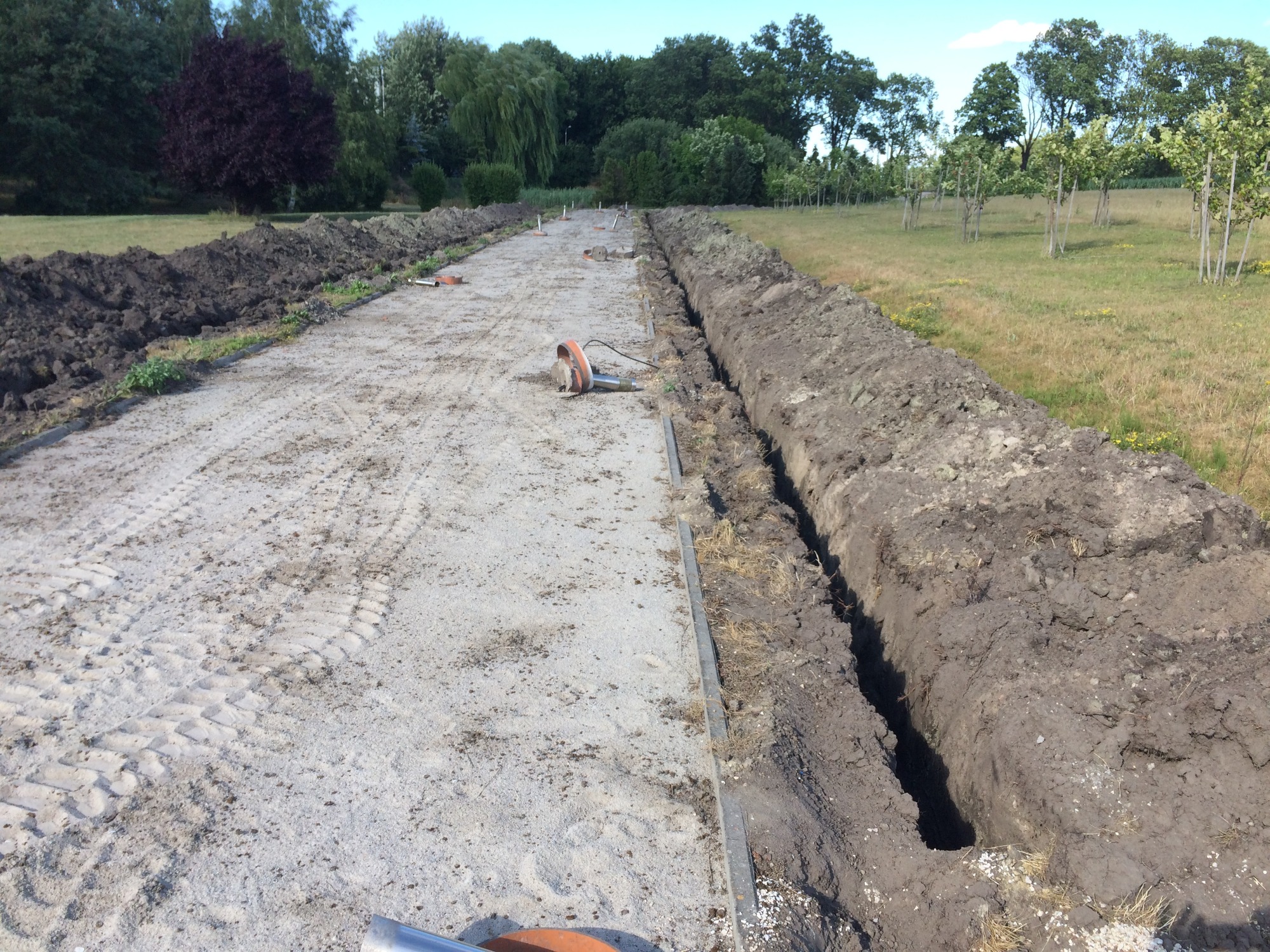 Wykopany rów w ziemi wzdłuż drogi gruntowej, z hałdami ziemi po obu stronach, widoczne narzędzia i ślady opon, w tle drzewa i trawnik.