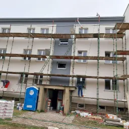 Wielopiętrowy budynek w trakcie odświeżania elewacji; robotnicy na rusztowaniu malują ściany na biało i szaro, widoczna przenośna toaleta na trawniku.