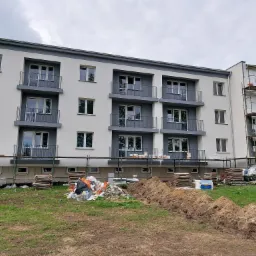Nowoczesny budynek wielorodzinny z jasną elewacją i szarymi balkonami, część budynku w trakcie ocieplania, widoczne rusztowania i materiały budowlane, ziemia rozkopana.