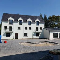 Nowoczesny, trzypiętrowy budynek z białymi ścianami i czarnym dachem, widoczny w słoneczny dzień, z elementami placu budowy: paleta, cegły, materiały budowlane przykryte plandeką.