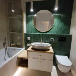 Nowoczesna łazienka z zieloną ścianą z płytek, okrągłym lustrem w złotej ramie, białą umywalką nablatową i podświetlaną szafką pod umywalką, widoczna toaleta i wanna za szklaną ścianką.