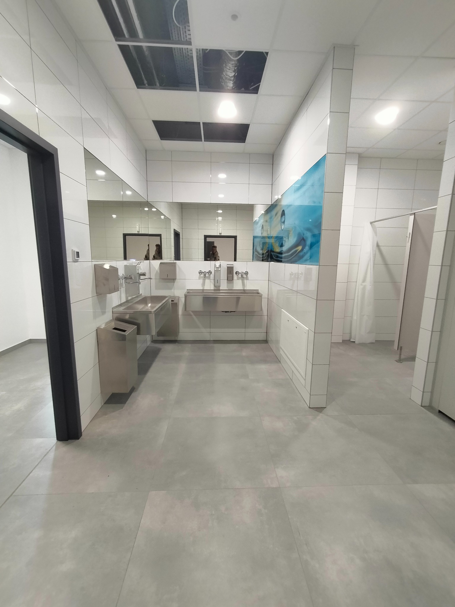 Nowoczesna, minimalistyczna łazienka publiczna z szarymi płytkami podłogowymi i białymi płytkami ściennymi, stalowymi umywalkami i lustrami, uzupełniona o panel dekoracyjny z motywem wodnym.