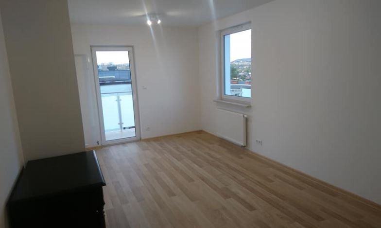 Jasne, puste pomieszczenie z drewnianą podłogą, białymi ścianami, oknem z widokiem na miasto i drzwiami balkonowymi, widoczna komoda.
