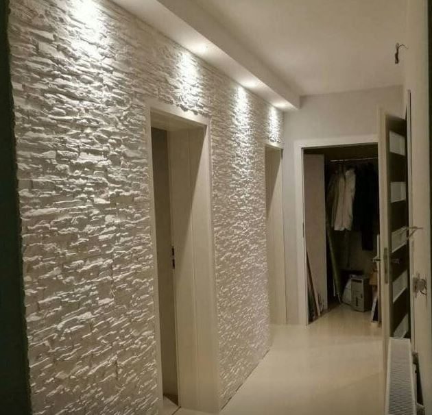 Korytarz z dekoracyjną ścianą pokrytą białym kamieniem imitującym łupanek, oświetlony punktowo, z widocznymi drzwiami i wejściem do garderoby.