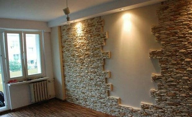Fragment ściany wewnętrznej z dekoracyjnym, częściowo ułożonym kamieniem elewacyjnym, oświetlonym punktowo od góry, z widocznym parkietem i grzejnikiem pod oknem.