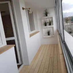 Jasne, surowe deski podłogowe na balkonie z białymi ścianami, drewnianym parapetem, dekoracyjną półką na rośliny i szklanymi panelami balkonowymi.