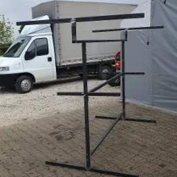 Surowa, metalowa konstrukcja z profilów, prawdopodobnie stojak lub element większej konstrukcji, na zewnątrz, z widocznym samochodem dostawczym i fragmentem namiotu w tle.