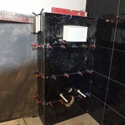 Usługi remontowe - wykończenia wnętrz Daniel Budner - Częściowo wykończona ściana łazienki z czarnymi, błyszczącymi płytkami, z widocznymi krzyżykami dystansowymi, poziomica na górze, rurami i otwartym otworem rewizyjnym.