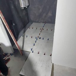 Usługi remontowe - wykończenia wnętrz Daniel Budner - Układanie jasnych płytek podłogowych w łazience, widoczne krzyżyki dystansowe, przygotowane podłoże na ścianach, grzejnik drabinkowy i fragment nogi glazurnika.