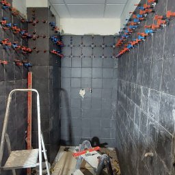 Usługi remontowe - wykończenia wnętrz Daniel Budner - Ściany łazienki w trakcie układania ciemnych płytek z użyciem systemu poziomowania, widoczne dwie drabiny i narzędzia.