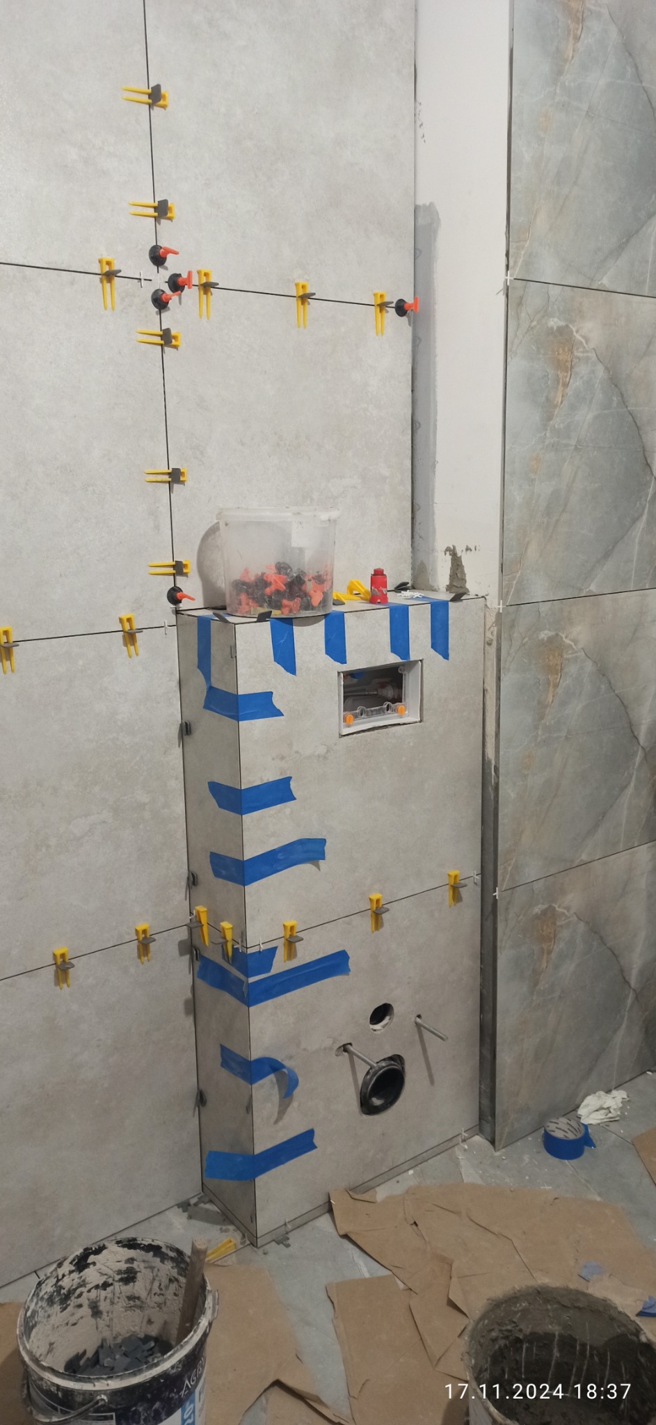 Montaż płytek ceramicznych w łazience, widoczne poziomowanie z użyciem klipsów i klinów, obudowa stelaża WC zabezpieczona niebieską taśmą malarską, wiadro z zaprawą na podłodze.