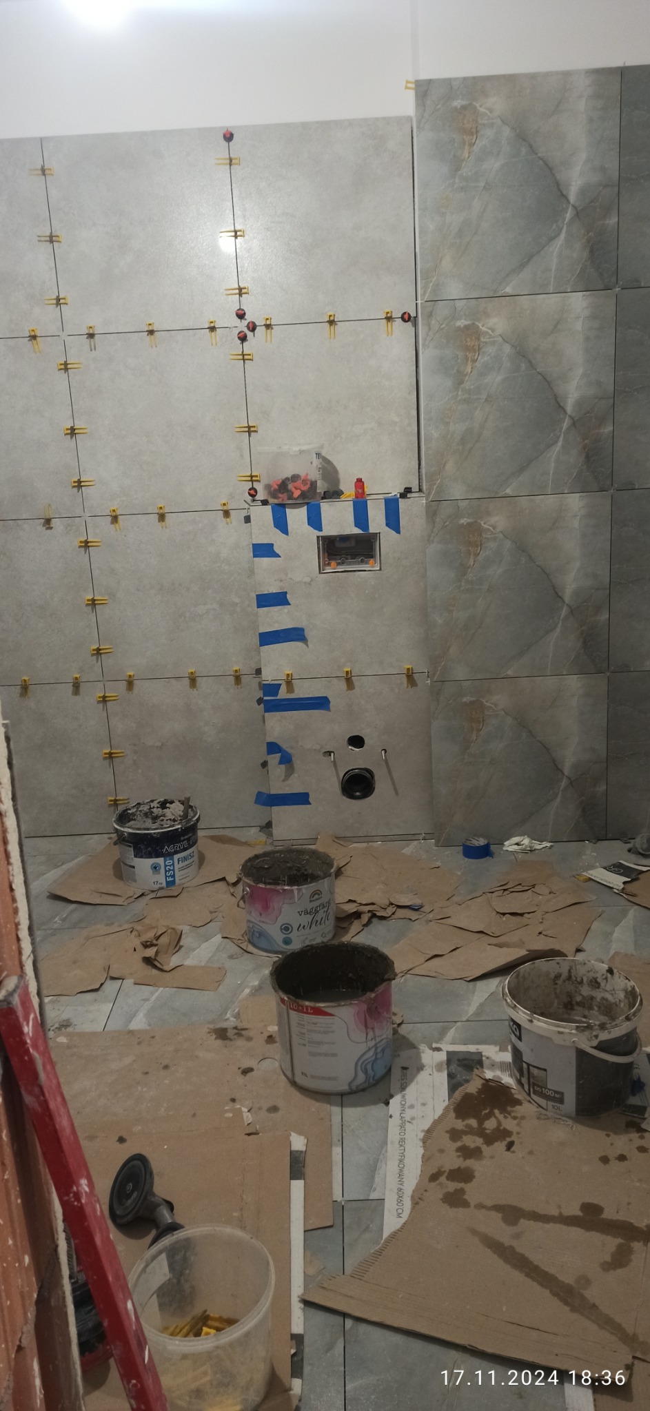 Łazienka w trakcie prac: częściowo ułożone szare płytki z marmurkowym wzorem, krzyżyki dystansowe, puszka podtynkowa zabezpieczona taśmą, widoczne wiaderka z zaprawą i narzędzia na podłodze...