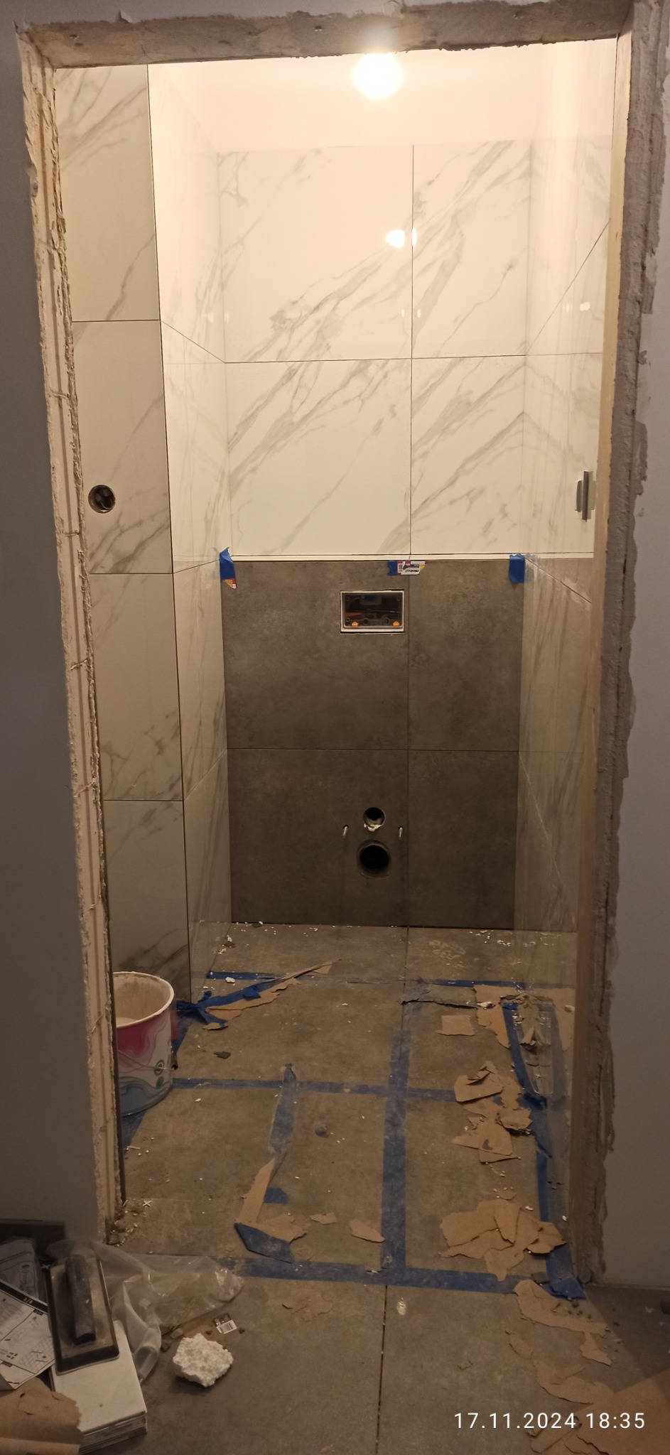 Łazienka w trakcie remontu: ściany wyłożone białymi i szarymi płytkami, widoczne otwory na instalacje, podłoga z resztkami kartonów i taśmą malarską, surowe wykończenie ościeżnicy.