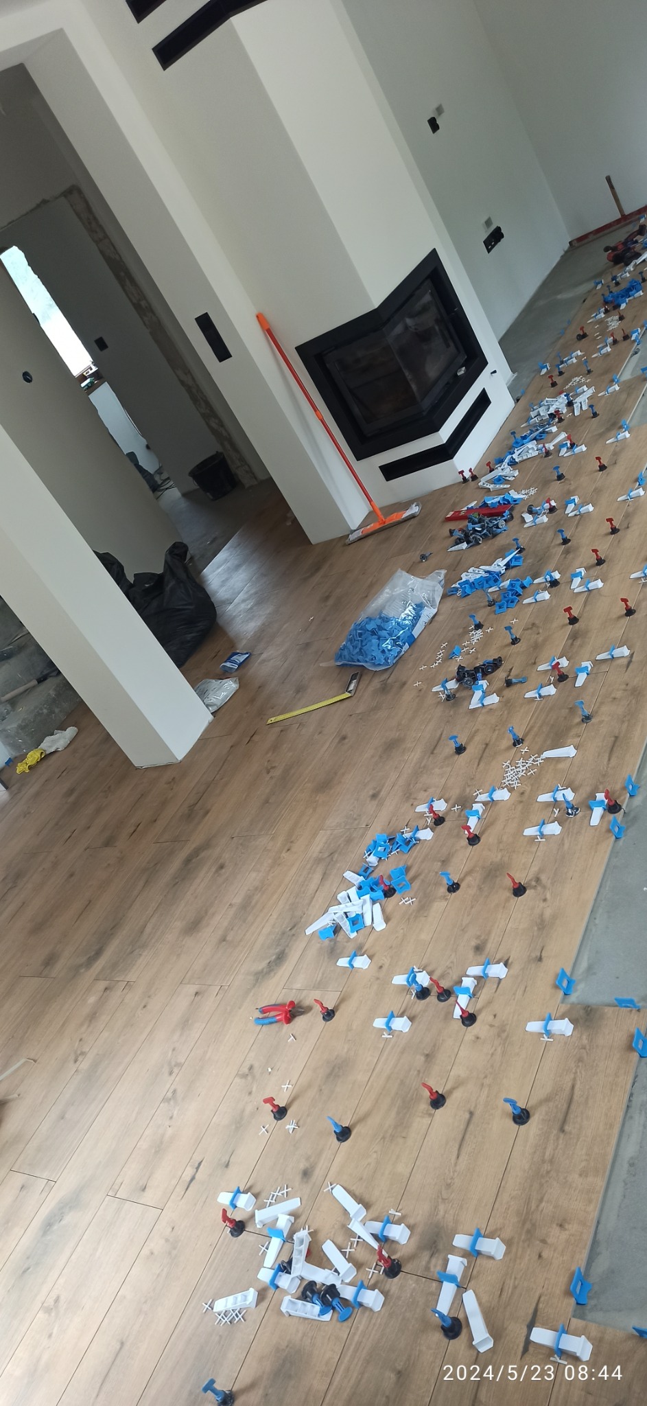 Ułożone płytki podłogowe imitujące drewno, z systemem poziomowania płytek (klipsy, kliny i szczypce), widok z góry, w tle kominek i przygotowane pomieszczenie.