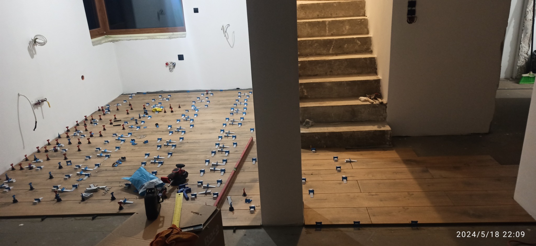 Układanie płytek podłogowych imitujących drewno z użyciem systemu poziomowania, widoczne narzędzia, poziomica i paczka z akcesoriami do układania płytek.