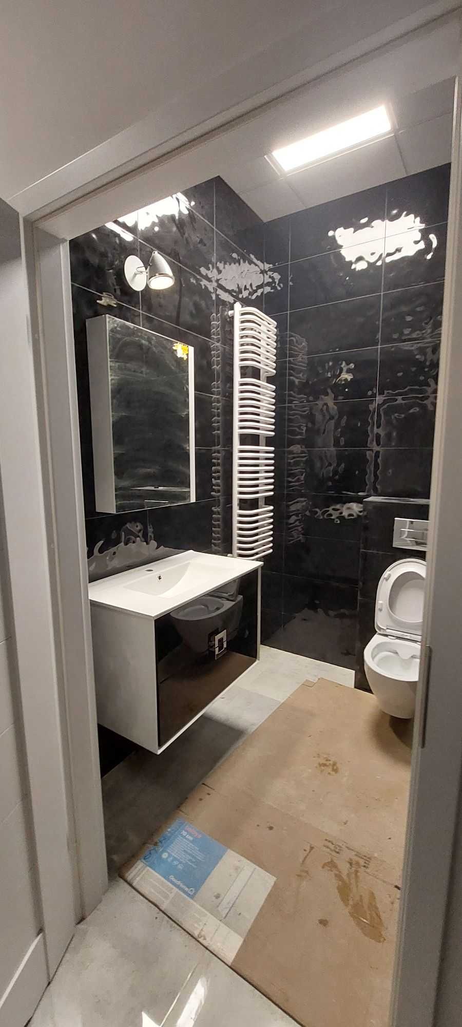 Ciemna łazienka w trakcie remontu: czarne płytki z efektem przetarć, biała umywalka z szafką, grzejnik drabinkowy i toaleta. Podłoga zabezpieczona kartonem, widoczne opakowanie płytek.