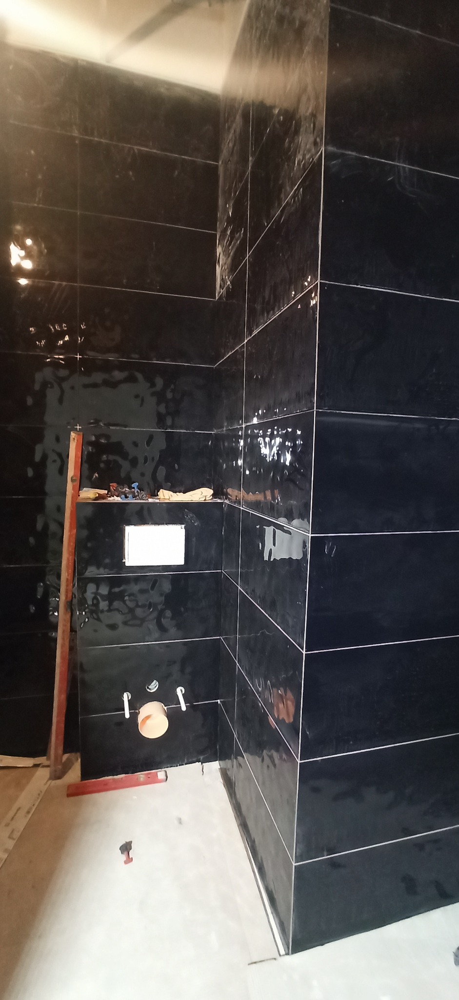 Łazienka w trakcie remontu, ściany wyłożone błyszczącymi, czarnymi płytkami, widoczne elementy instalacji wodnej, poziomica i narzędzia na podłodze.