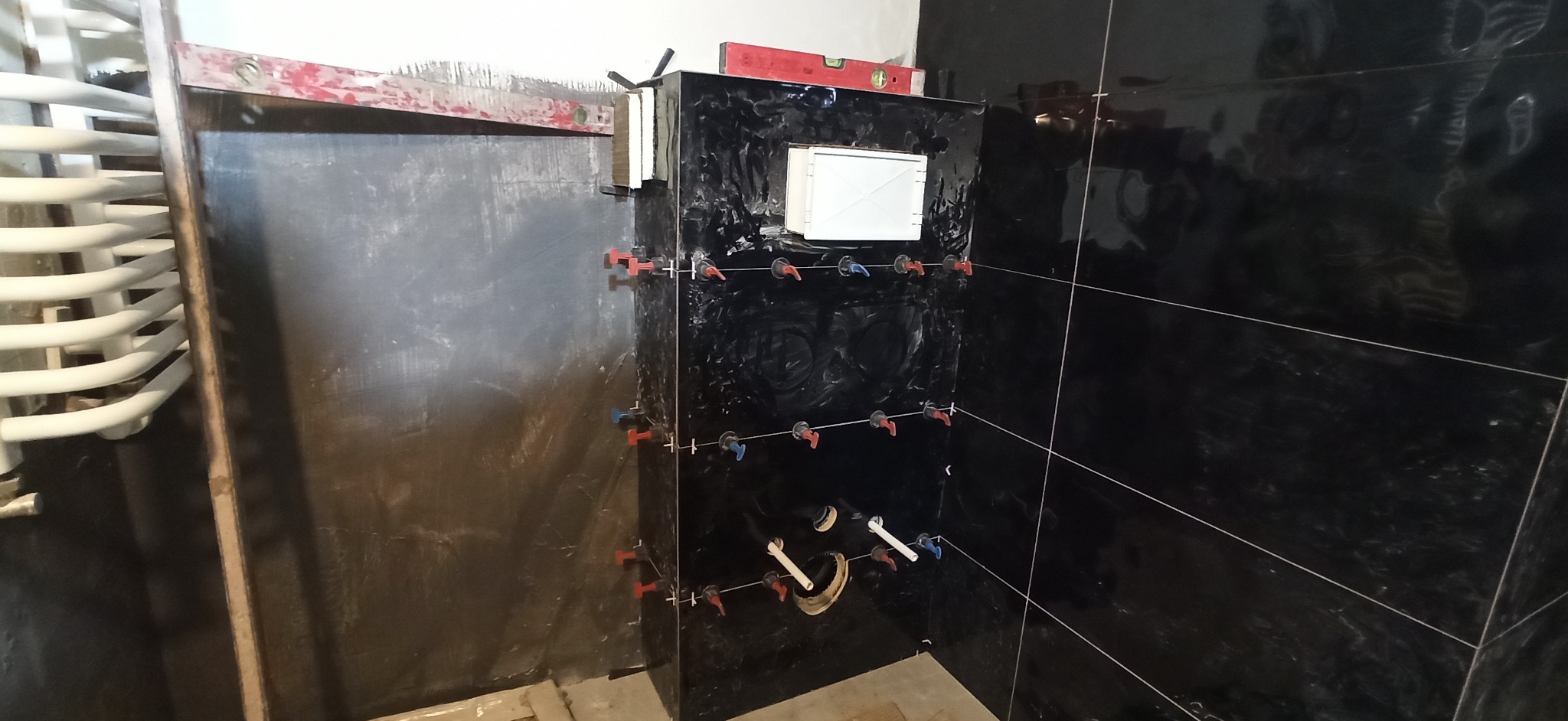 Częściowo wykończona ściana łazienki z czarnymi, błyszczącymi płytkami, z widocznymi krzyżykami dystansowymi, poziomica na górze, rurami i otwartym otworem rewizyjnym.