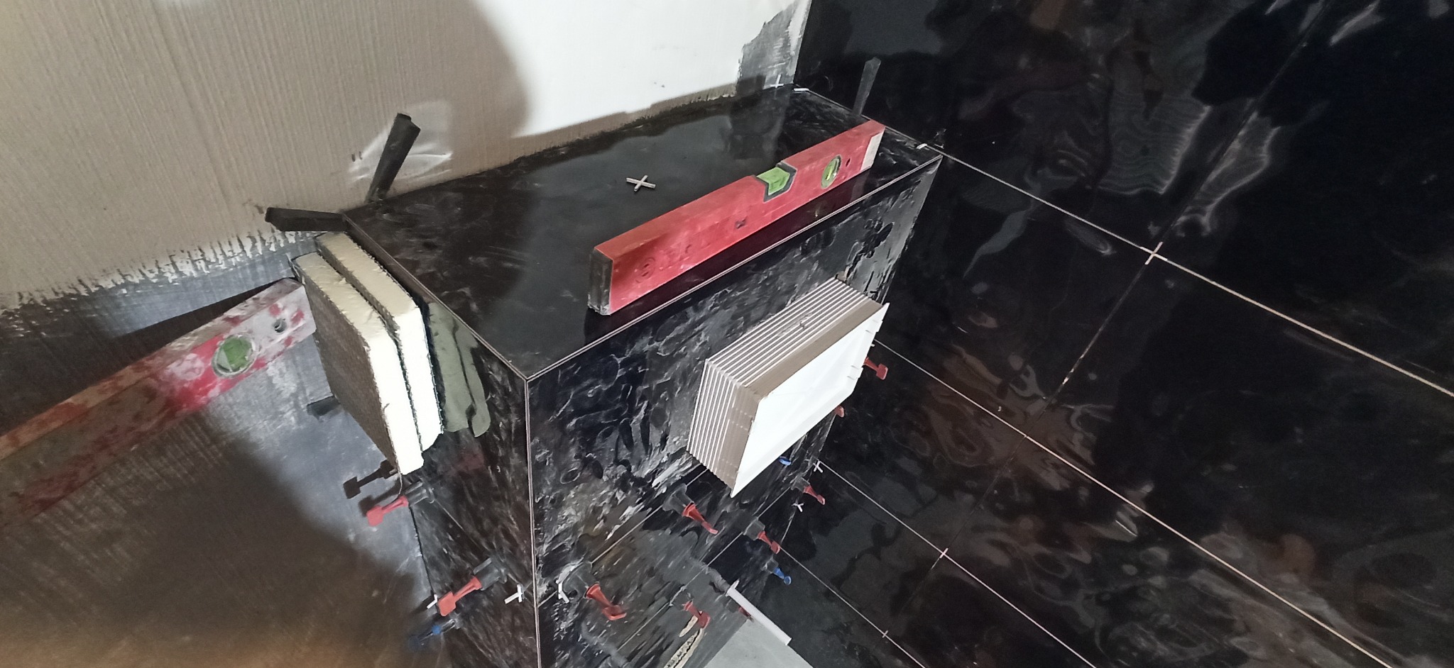 Montaż czarnych płytek imitujących marmur na ścianie, z użyciem poziomicy, krzyżyków dystansowych i klipsów poziomujących; widoczne zapasowe płytki.