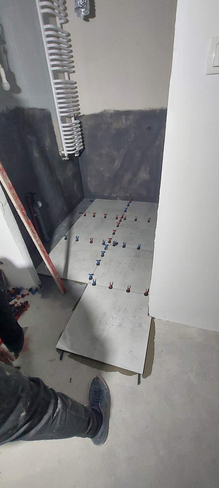 Układanie jasnych płytek podłogowych w łazience, widoczne krzyżyki dystansowe, przygotowane podłoże na ścianach, grzejnik drabinkowy i fragment nogi glazurnika.