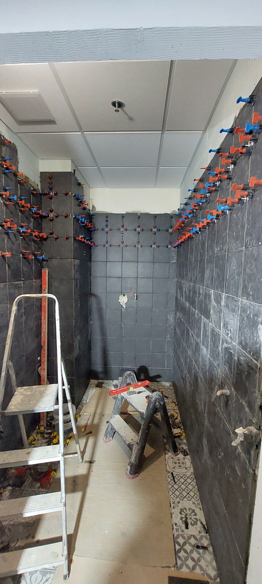 Ściany łazienki w trakcie układania ciemnych płytek z użyciem systemu poziomowania, widoczne dwie drabiny i narzędzia.