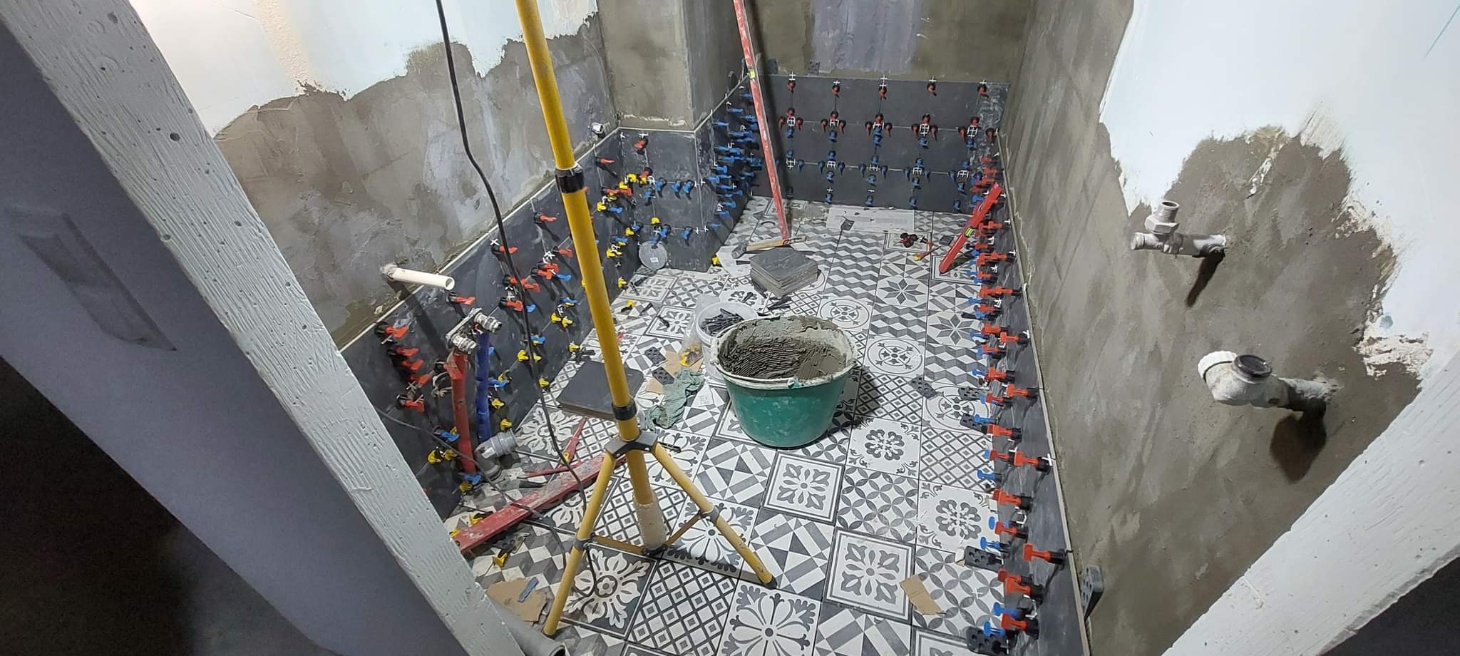 Wnętrze łazienki w trakcie układania płytek podłogowych w czarno-biały wzór oraz ciemnych płytek ściennych z użyciem klipsów poziomujących; widoczny kubeł z zaprawą, poziomica oraz reflektor...