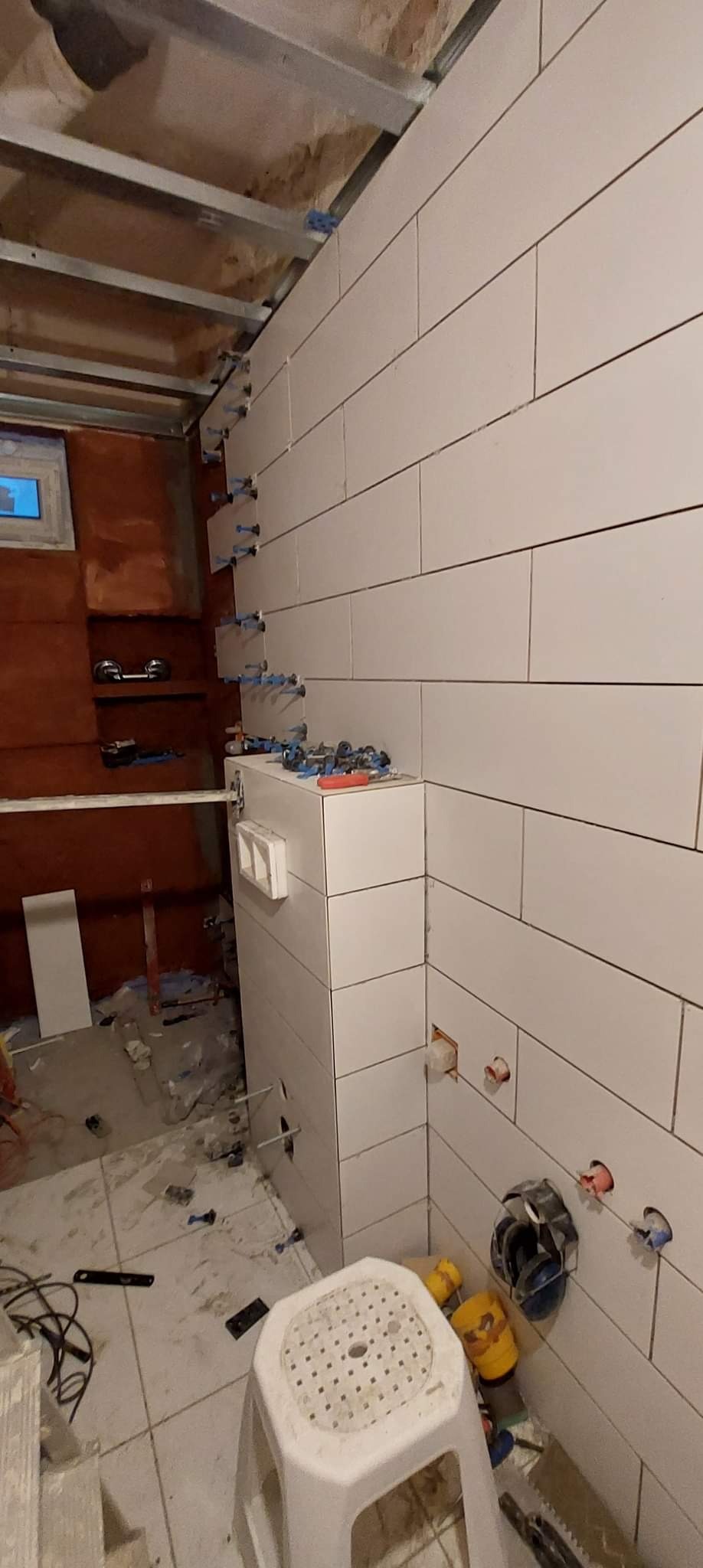 Białe, prostokątne płytki ceramiczne ułożone poziomo na ścianie łazienki, z widocznymi niebieskimi krzyżykami dystansowymi i poziomnicą laserową, w trakcie remontu, z widoczną konstrukcją sufitu...
