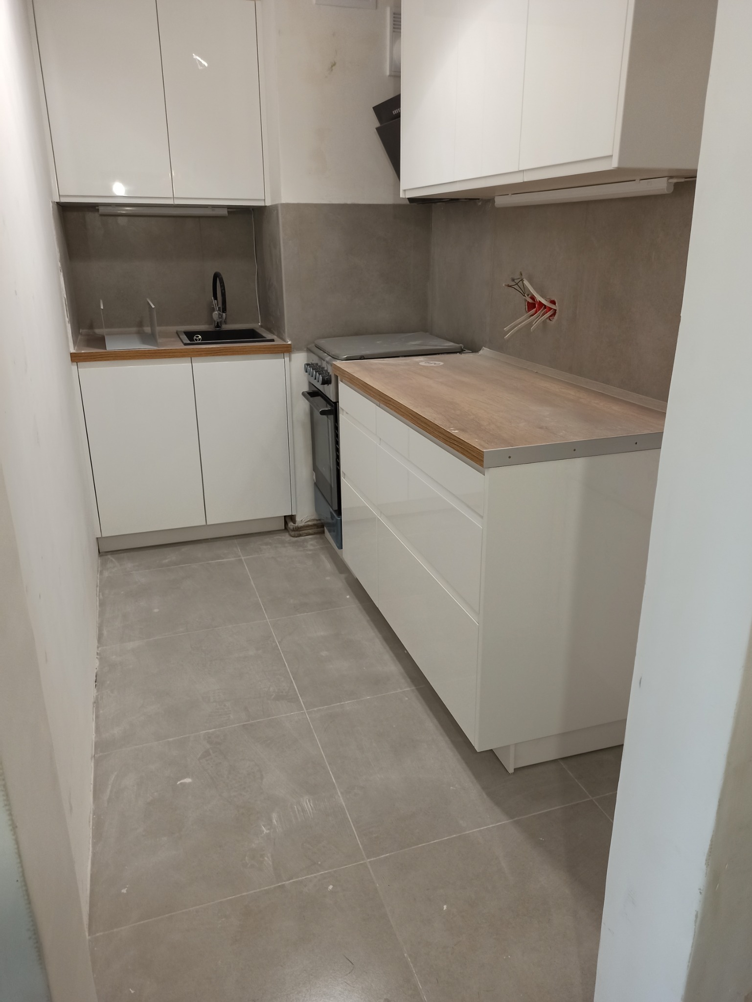 Wnętrze kuchni w trakcie prac wykończeniowych: białe, błyszczące szafki kuchenne, szare płytki podłogowe, widoczne kable elektryczne w ścianie, blat roboczy z drewnopodobnym wykończeniem.