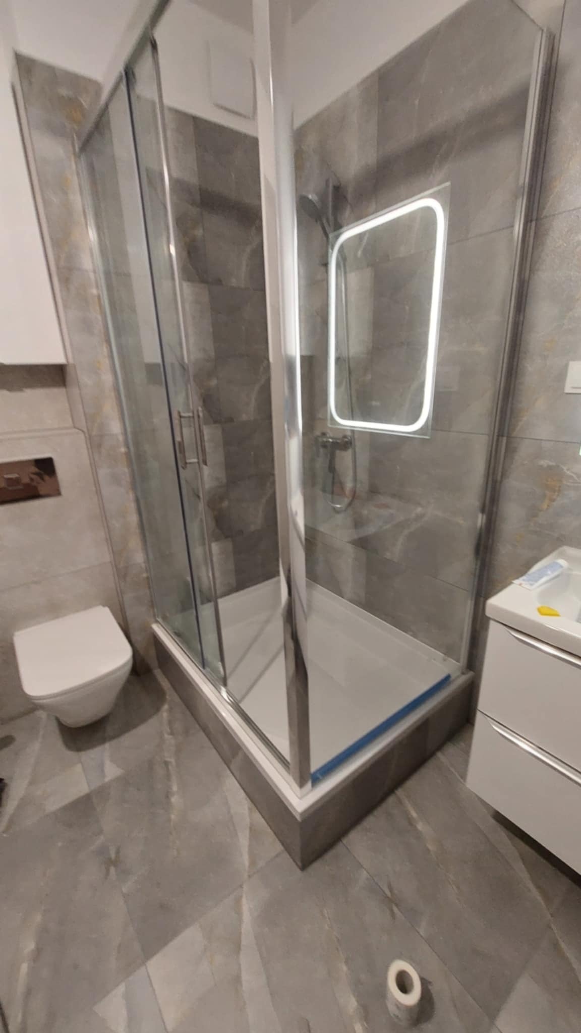 Nowoczesna kabina prysznicowa z podświetlanym lustrem w łazience wykończonej szarymi płytkami imitującymi marmur. Widoczna toaleta i umywalka z szafką.