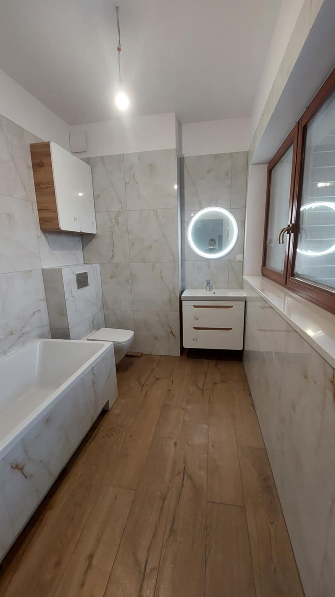 Jasna łazienka po remoncie: drewnopodobna podłoga, marmurowe płytki, biała wanna i szafka z okrągłym lustrem z podświetleniem LED. Widok na okno z brązową ramą.