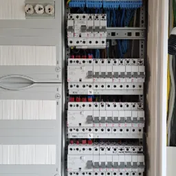 Otwarta skrzynka elektryczna z bezpiecznikami, wyłącznikami różnicowoprądowymi i listwami zaciskowymi, widoczne okablowanie i elementy montażowe.