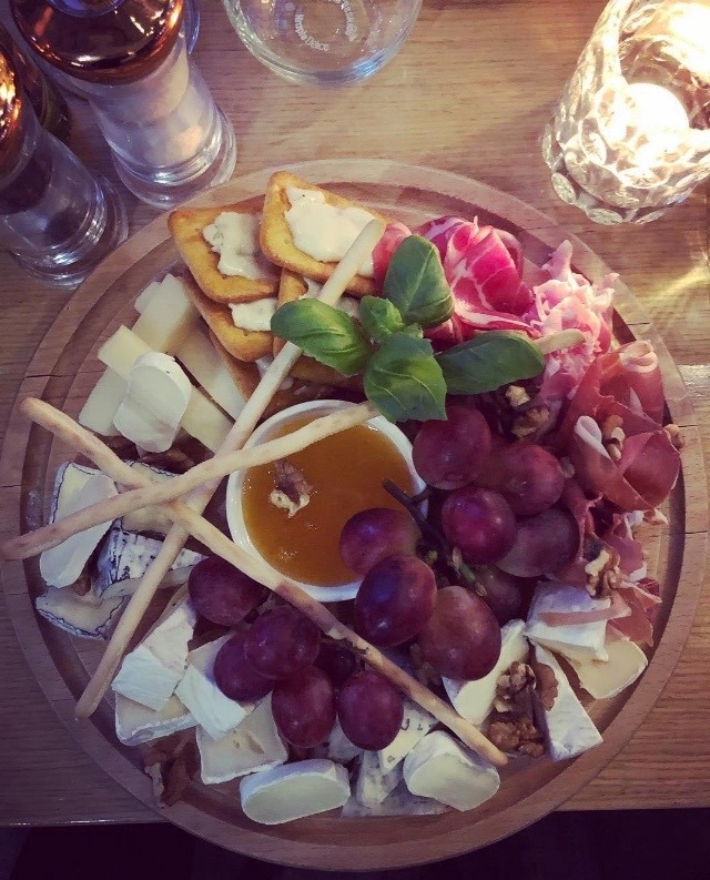 Deska serów z winogronami, szynką parmeńską, krakersami i dipem miodowym, udekorowana bazylią, podana na drewnianej tacy, widok z góry.