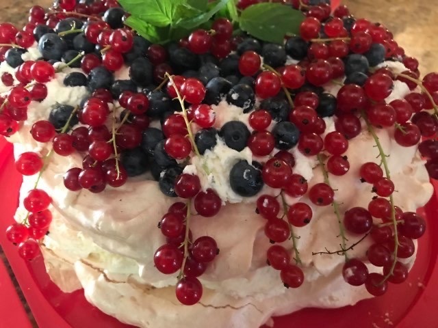 Dwupoziomowy tort bezowy udekorowany bitą śmietaną, świeżymi borówkami, pękami czerwonej porzeczki i listkami bazylii, umieszczony na czerwonym talerzu.