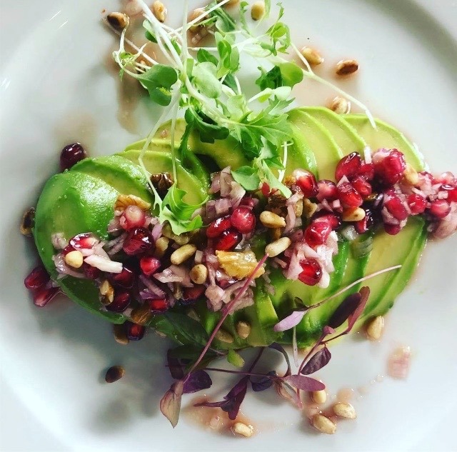 Awokado w plastrach udekorowane sałatką z granatu, orzechów i mikro zieleniny, podane na białym talerzu.