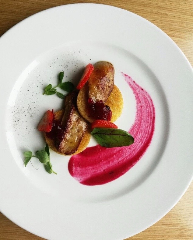 Elegancko podane foie gras na blinach z sosem owocowym i truskawkami, dekorowane świeżymi ziołami na białym talerzu.
