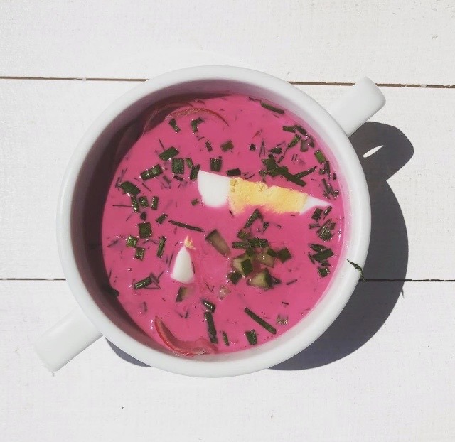 Chłodnik litewski w białej miseczce z dwoma uchwytami, udekorowany zieleniną i jajkiem, na białym drewnianym tle.