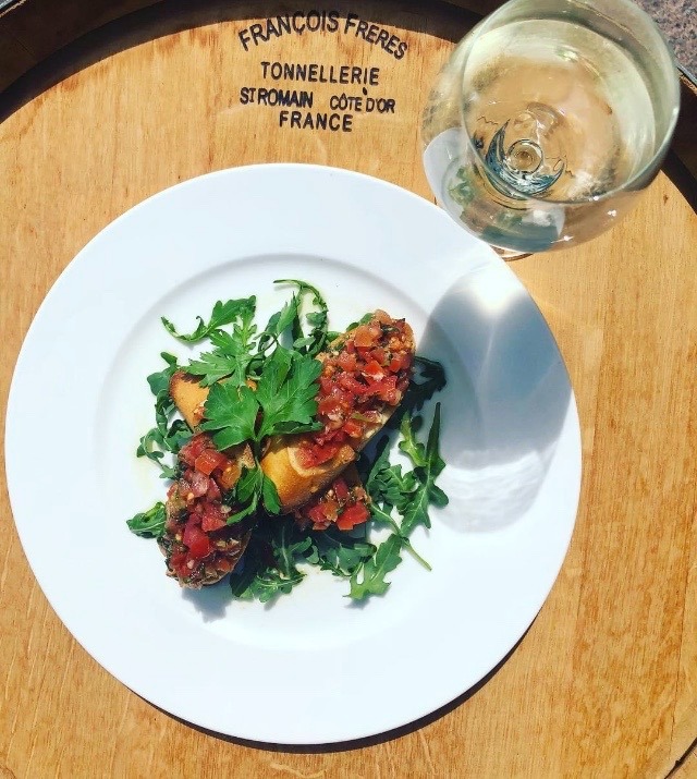 Bruschetta z pomidorami i bazylią na białym talerzu, udekorowana rukolą i natką pietruszki, obok kieliszek białego wina, całość na drewnianej beczce z francuskim napisem.