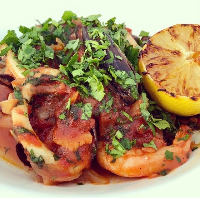 Aromatyczne owoce morza w sosie pomidorowym, posypane świeżą pietruszką i podane z grillowaną cytryną na białym talerzu.